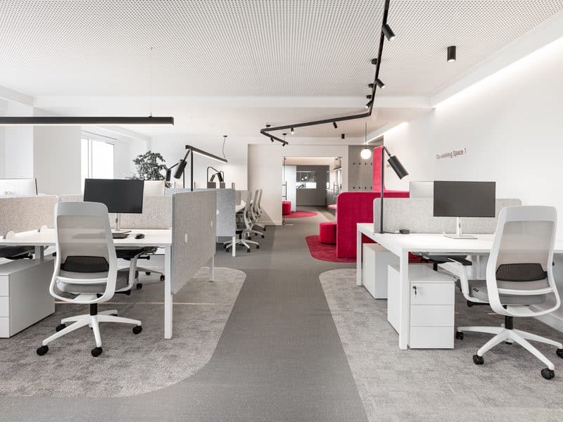 Open kantoor met witte bureaus en bureaustoelen op een grijze vloer, met een rode zithoek achterin