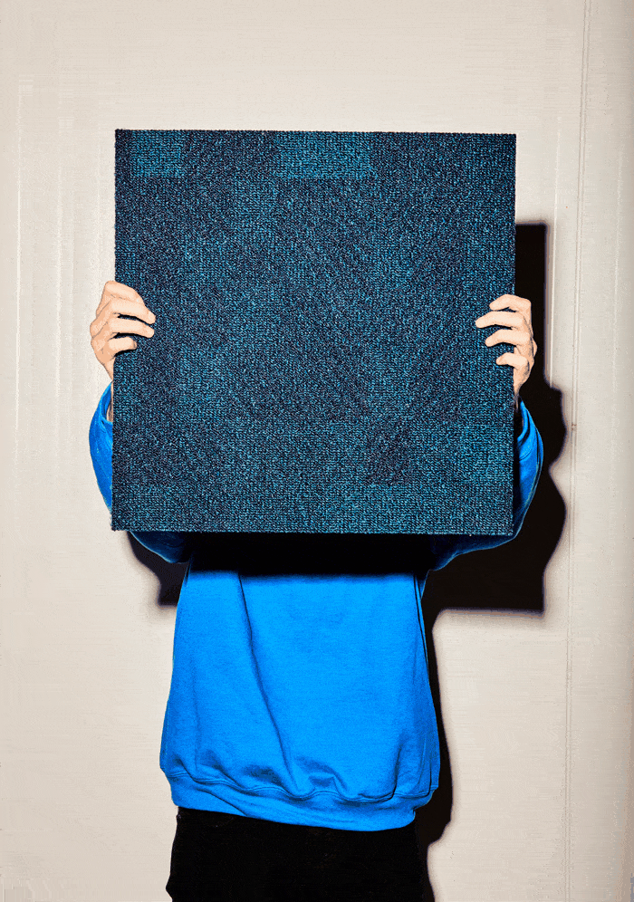 Iemand houdt achter elkaar drie tapijttegels voor zich: een blauw vierkant, een geelgroene ruit en een oranje zeshoek.