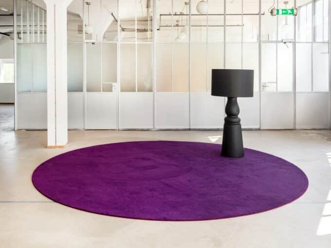 Rond paars karpet op een lichte vloer in een open ruimte, met een zwarte vloerlamp erop